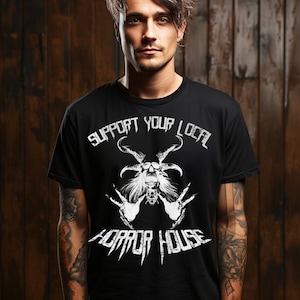 Peut inclure: Un t-shirt noir avec un imprimé blanc d'un crâne avec des cornes et le texte "Support Your Local Horror House".
