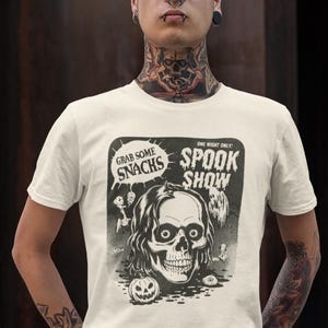 May include: Cream-coloured t-shirt with a black and white graphic. The graphic includes a skull, a pumpkin, and the words "Grab Some Snacks" and "Spook Show." The t-shirt has a crew neck.