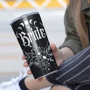 K&ouml;nnte beinhalten: Schwarzer und wei&szlig;er Becher mit dem Text "The Bride" und einem floralen Design. Der Becher hat einen schwarzen Hintergrund und wei&szlig;en Text und Design.