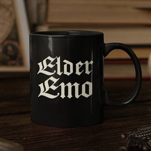 May include: Black ceramic mug with white text that reads "Elder Emo". The mug is sitting on a wooden table with a book and a skull in the background.