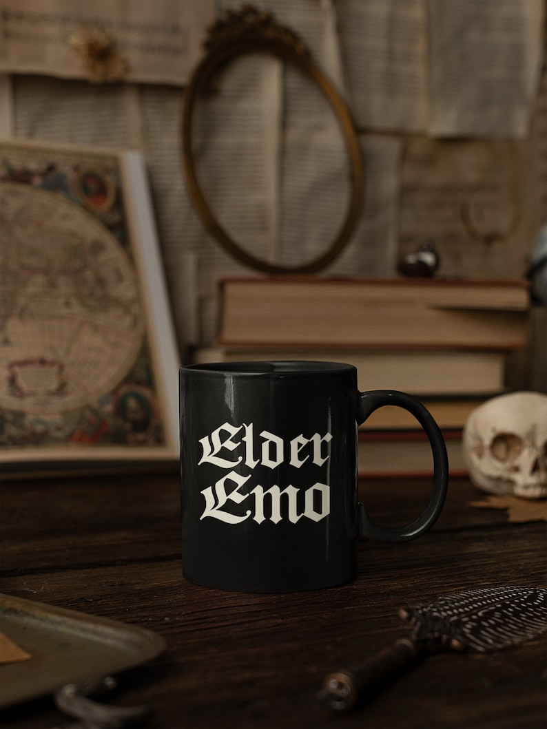 May include: Black ceramic mug with white text that reads "Elder Emo". The mug is sitting on a wooden table with a book and a skull in the background.