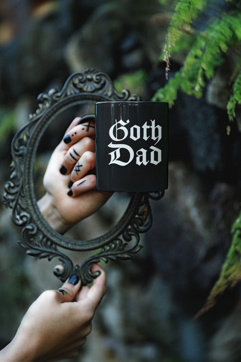 K&ouml;nnte beinhalten: Eine schwarze Keramiktasse mit dem Text "Goth Dad" in wei&szlig;en gotischen Buchstaben. Die Tasse wird von einer Hand mit schwarzem Nagellack und Tattoos gehalten.