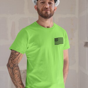 May include: A man wearing a white hard hat and a bright green t-shirt with a small black American flag on the left chest. He is standing in front of a white wall.