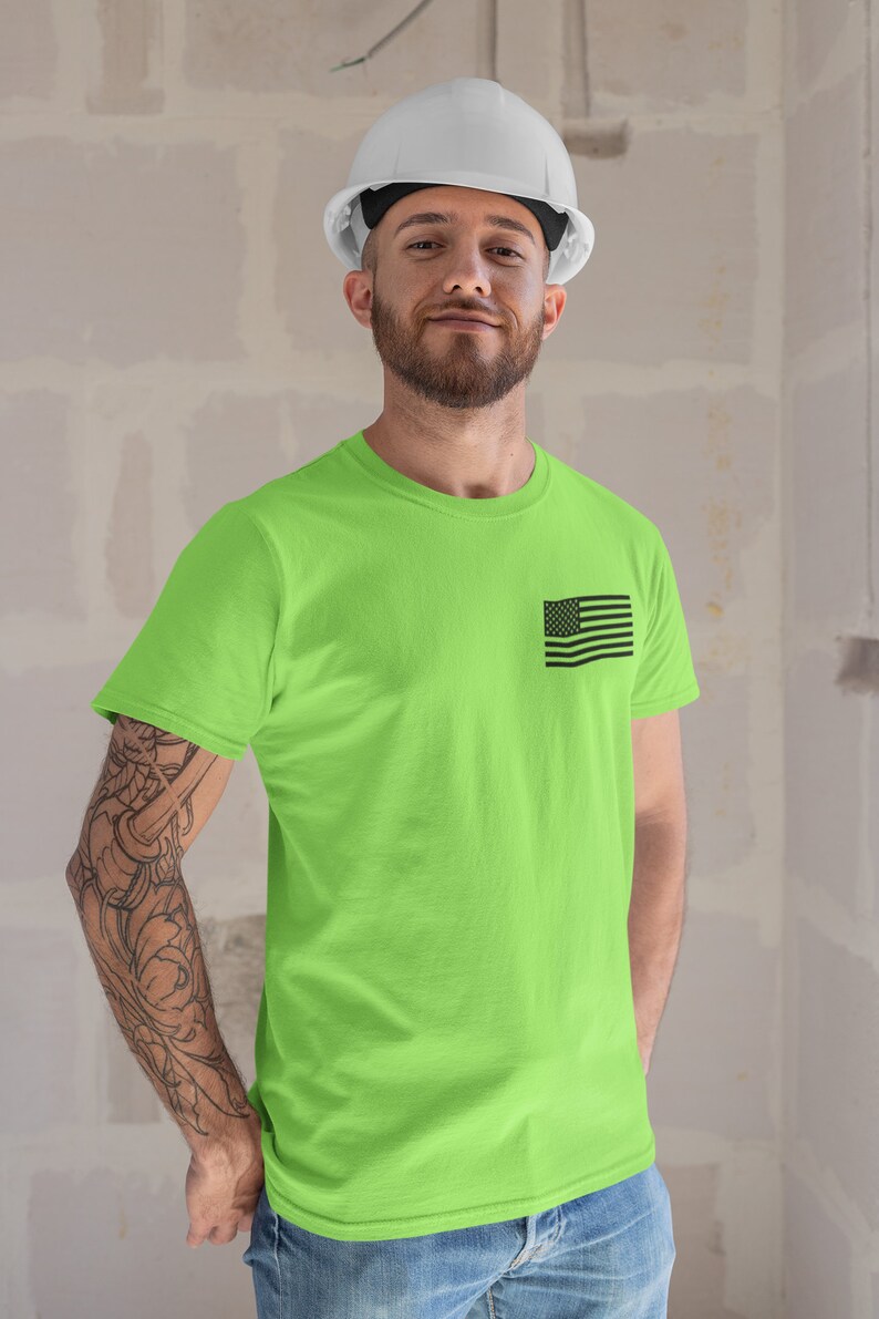 May include: A man wearing a white hard hat and a bright green t-shirt with a small black American flag on the left chest. He is standing in front of a white wall.