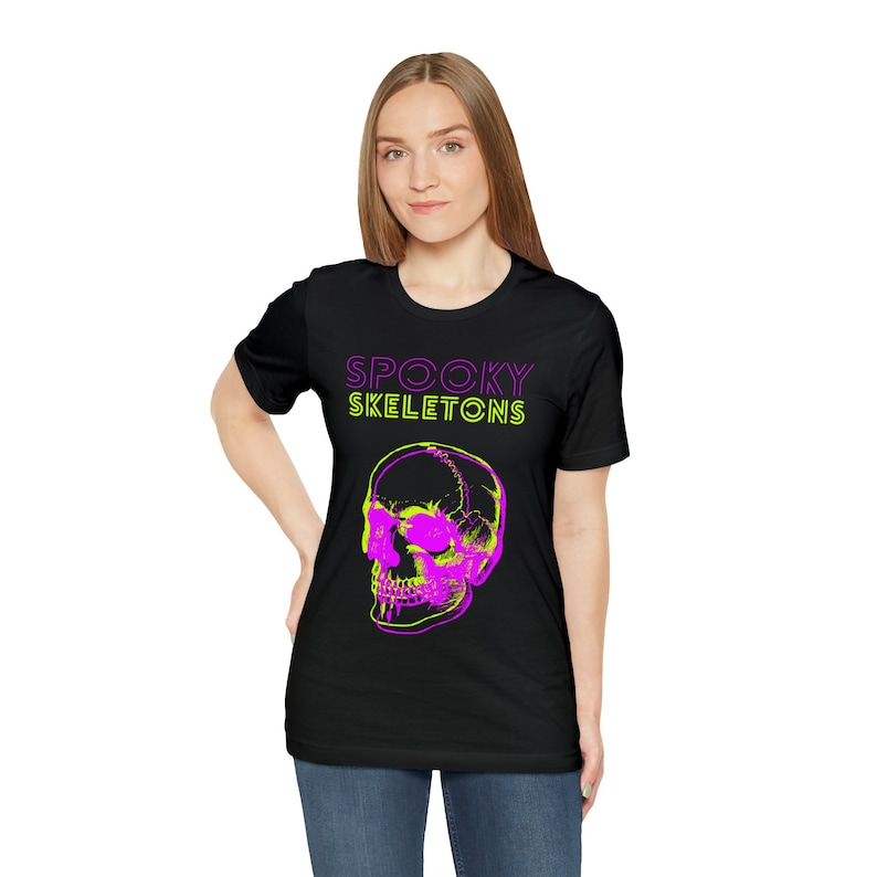 May include: Black t-shirt featuring a neon pink and green skull graphic. The text "SPOOKY SKELETONS" is above the skull in matching neon colors. The shirt is a crew neck style.