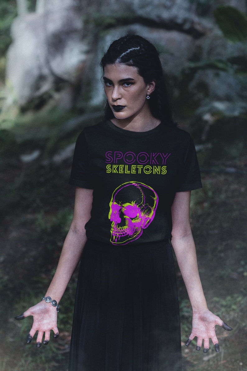 May include: Black t-shirt with the text "SPOOKY SKELETONS" in neon green and purple. The shirt features a neon green and purple skull graphic. The model is wearing a black skirt and has dark makeup.