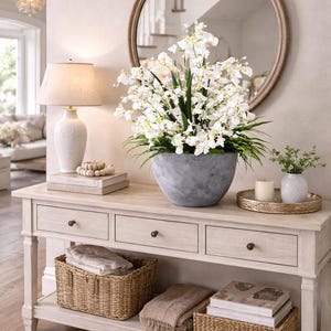 May include: A pale wooden console table with three drawers and a lower shelf. It features a large grey stone-effect bowl filled with white flowers, a lamp, and a round mirror. Wicker baskets and a tray with candles add to the decor.
