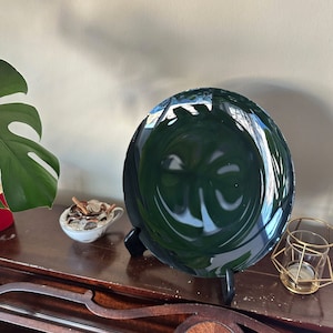K&ouml;nnte beinhalten: Ein dunkelgr&uuml;ner Glasteller mit reflektierender Oberfl&auml;che wird auf einem schwarzen St&auml;nder pr&auml;sentiert. Der Teller ist rund und hat eine leichte W&ouml;lbung. Der Teller steht auf einer Holzoberfl&auml;che.