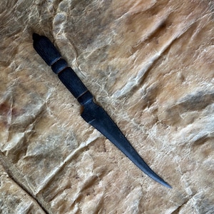 May include: A black, handcrafted dagger with a textured handle and a sharp, pointed blade. The handle features a series of carved rings. The dagger is displayed against a textured, light brown background.