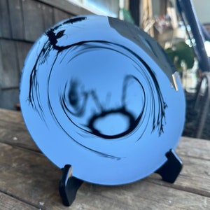 K&ouml;nnte beinhalten: Ein runder, blauer, reflektierender Spiegel mit einem schwarzen Wirbelmuster. Der Spiegel steht auf einem schwarzen St&auml;nder.