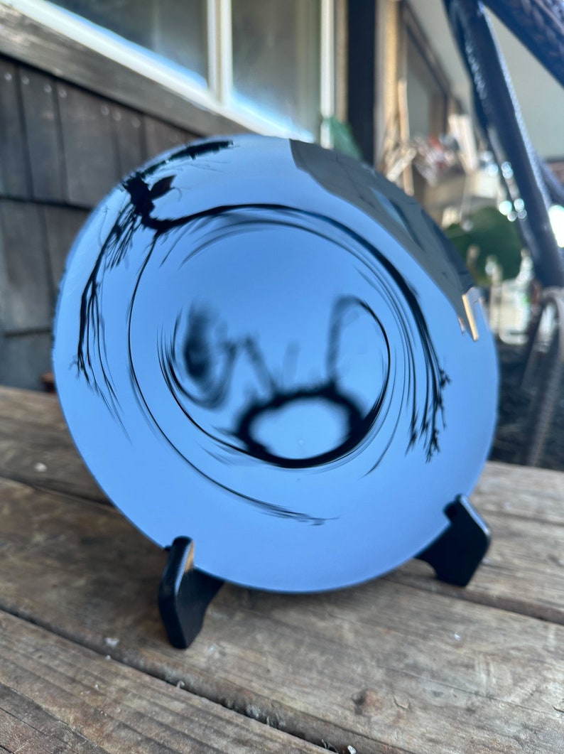 K&ouml;nnte beinhalten: Ein runder, blauer, reflektierender Spiegel mit einem schwarzen Wirbelmuster. Der Spiegel steht auf einem schwarzen St&auml;nder.
