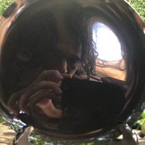 K&ouml;nnte beinhalten: Ein schwarzer, runder, reflektierender Teller mit dem Spiegelbild einer Person. Die Person h&auml;lt eine Kamera und macht ein Selfie. Der Teller steht auf einer Holzoberfl&auml;che.