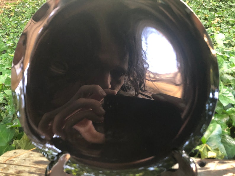 K&ouml;nnte beinhalten: Ein schwarzer, runder, reflektierender Teller mit dem Spiegelbild einer Person. Die Person h&auml;lt eine Kamera und macht ein Selfie. Der Teller steht auf einer Holzoberfl&auml;che.