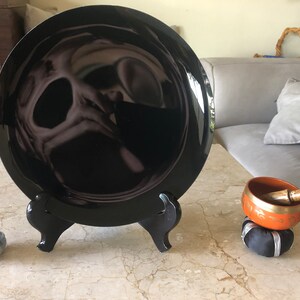 K&ouml;nnte beinhalten: Ein schwarzer Keramikteller mit reflektierender Oberfl&auml;che wird auf einem schwarzen St&auml;nder pr&auml;sentiert. Eine kleine, orangefarbene Klangschale mit einem Holzschl&auml;gel steht auf einem schwarzen Kissen.