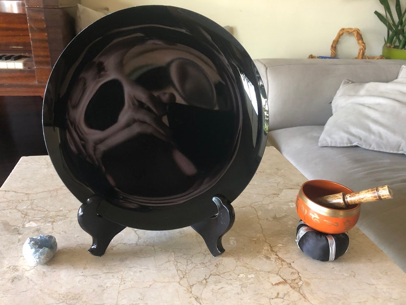 K&ouml;nnte beinhalten: Ein schwarzer Keramikteller mit reflektierender Oberfl&auml;che wird auf einem schwarzen St&auml;nder pr&auml;sentiert. Eine kleine, orangefarbene Klangschale mit einem Holzschl&auml;gel steht auf einem schwarzen Kissen.