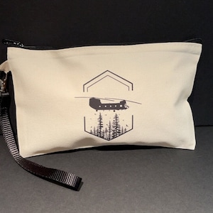 May include: A beige canvas zippered pouch with a black strap. The pouch features a black and white graphic of a helicopter flying over a forest. The graphic is inside a hexagonal frame.