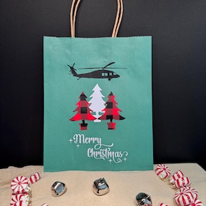 May include: A green paper gift bag with a black silhouette of a helicopter and two red and black plaid Christmas trees. The bag has a brown paper handle and the text "Merry Christmas" is printed on the front.