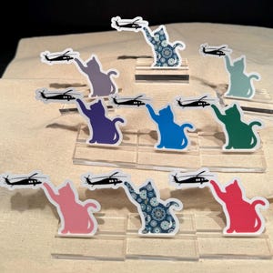 May include: Ten colorful cat figurines holding miniature black helicopters. Each figurine is on a clear acrylic base. The cats are in various poses, including sitting, standing, and reaching for the helicopters. The cats are in the following colors: blue, green, pink, red, purple, teal, and two with a paisley pattern.