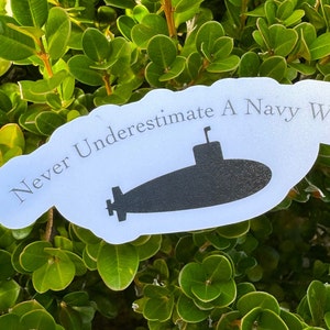 May include: White sticker with the text "Never Underestimate A Navy Wife" and a black silhouette of a submarine.