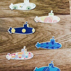 May include: A collection of colorful submarine-shaped stickers. Designs include floral patterns, citrus fruit slices, polka dots, turtles, and tropical leaves. The stickers are arranged on a wooden surface.