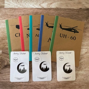 May include: Three brown notebooks with green, blue, and red spines. Each notebook has a black silhouette of a helicopter on the front with the text "Army Sticker" and the name of the helicopter model.  The notebooks are accompanied by three white sticker sheets with the text "Army Sticker" and a black silhouette of a helicopter in a forest.