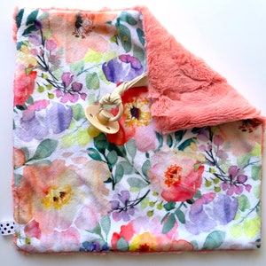 May include: A soft, pink and white floral baby blanket with a plush pink backing. The blanket features a watercolour floral design with a dummy attached.