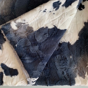 May include: A soft, plush blanket with a white and gray cow print design. The blanket is reversible, with a solid dark blue side.