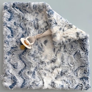 May include: A soft, plush baby blanket in shades of blue and white, with a pacifier attached. The blanket has a textured, faux fur appearance. The pacifier is white and tan, attached with a neutral-colored strap.