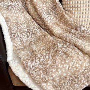 May include: A plush, faux fur blanket in shades of tan and cream, featuring a spotted pattern. The blanket is draped over a brown leather couch, with a white pillow with a textured surface visible in the background. The blanket's soft texture is visible.