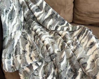 May include: A soft faux fur blanket in grey, white, and green tones, draped over a brown sofa. The blanket features a textured pattern reminiscent of an animal print. The material appears plush and inviting, suggesting warmth and comfort for the home.
