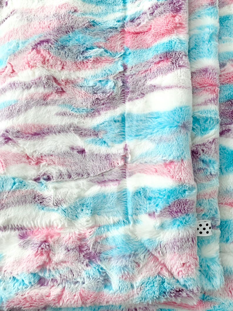 May include: A soft, plush blanket with a tie-dye pattern in shades of pink, blue, and purple. The blanket is made of faux fur and has a fluffy texture.