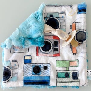 May include: A square baby security blanket featuring a white background with a colorful pattern of vintage cameras in blue, red, and green. The blanket has a textured light blue plush backing and includes a cream-colored pacifier clip attached.