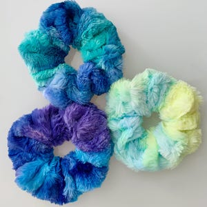 May include: Three fuzzy hair scrunchies in blue, green, and purple tie-dye patterns.