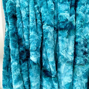 May include: A plush, textured blanket in shades of teal and navy blue. The fabric has a soft, fur-like appearance, with vertical folds creating a layered effect. The blanket is a solid color with darker blue accents.