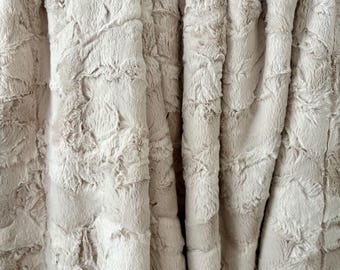 May include: A beige faux fur blanket with a textured, rippled surface. The blanket is folded, showing the soft, dense material. The neutral colour and tactile appearance suggest comfort and warmth, suitable for home decor.