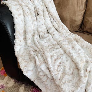 May include: A plush, cream-colored blanket with a textured, faux fur design. The blanket is draped over a black leather chair and a brown sofa, creating a cozy and inviting aesthetic. The blanket's soft texture and neutral color palette make it a versatile home decor item.