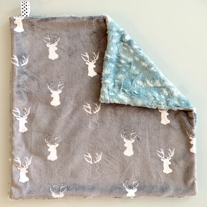 May include: A gray and blue baby blanket with a deer pattern. The front of the blanket is gray with white deer heads, and the back is a soft blue material. A small white and black polka dot ribbon is attached to the top corner.
