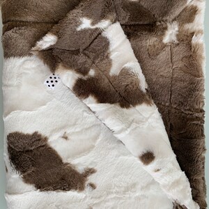 May include: A soft, brown and white faux fur throw blanket with a cowhide pattern. The blanket is folded in half and has a small white tag with black polka dots.
