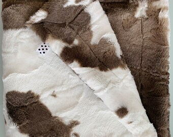 May include: A soft, brown and white faux fur throw blanket with a cowhide pattern. The blanket is folded in half and has a small white tag with black polka dots.