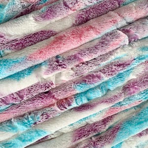 May include: A stack of soft, fluffy fabric in shades of pink, purple, and blue. The fabric has a tie-dye pattern and is perfect for crafting projects.
