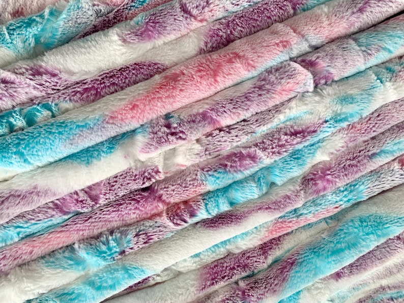 May include: A stack of soft, fluffy fabric in shades of pink, purple, and blue. The fabric has a tie-dye pattern and is perfect for crafting projects.