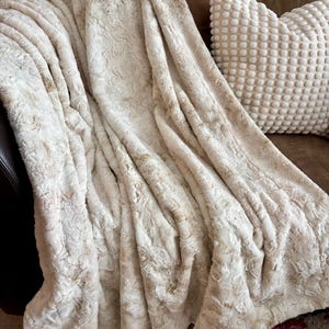 Puede incluir: Una manta afelpada de color crema con un patrón texturizado similar a flores. La manta está sobre un sofá marrón, con una almohada texturizada y una almohada marrón en el fondo. La manta parece suave y acogedora.