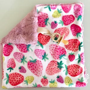 Op de afbeelding: Een vierkant baby-knuffeldoekje met een witte achtergrond en een herhalend patroon van roze en rode aardbeien met groene blaadjes. Het doekje heeft een zachte, getextureerde mauvekleurige pluche achterkant en een beige speenkoord.