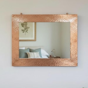 Pode incluir: Espelho quadrado cor de cobre com textura martelada, pendurado numa parede branca. O espelho reflete uma cena de quarto. Abaixo, uma consola de madeira com puxadores de latão e uma pequena suculenta num vaso.