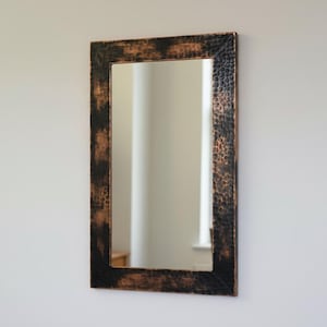 Pode incluir: Espelho de parede retangular com uma moldura texturizada em castanho escuro e preto. O espelho está pendurado numa parede branca. A moldura tem um aspeto desgastado e rústico, adicionando um toque de charme vintage à decoração.