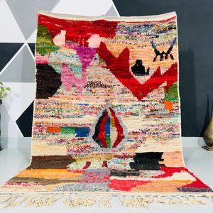 May include: A colorful Moroccan rug with a geometric pattern in shades of red, pink, blue, green, and black. The rug has a fringe border and is made of wool.