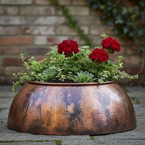 Op de afbeelding: Een koperen plantenbak met een gehamerde afwerking, gevuld met groene vetplanten en rode geraniums. De ronde, lage bak staat op een stenen ondergrond. Een bakstenen muur en groen zijn zichtbaar op de achtergrond. Het koper heeft een warme patina.