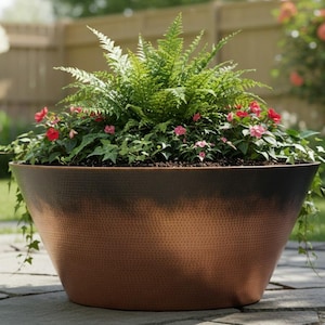 May include: A large, copper-coloured planter filled with greenery and small red and pink flowers. The planter has a textured surface and a wide, open design. The background shows a lush garden setting with a wooden fence and green grass.