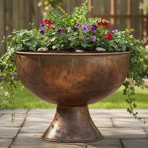 May include: A large, antique copper planter filled with vibrant green foliage and colourful red and purple flowers. The planter has a riveted design and sits on a pedestal base, ideal for outdoor decor.