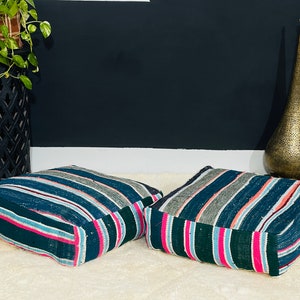 May include: Two square floor cushions with a striped pattern in shades of blue, green, pink, and grey. The cushions are placed on a cream-coloured rug. A black decorative plant stand and a gold metal vase are in the background.